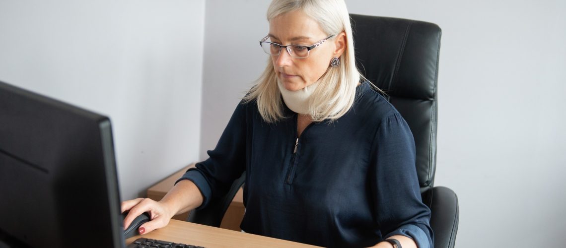 mature-woman-in-pain-wearing-a-surgical-collar-on-her-neck-working-on-computer-in-business-office_t20_EnkO81